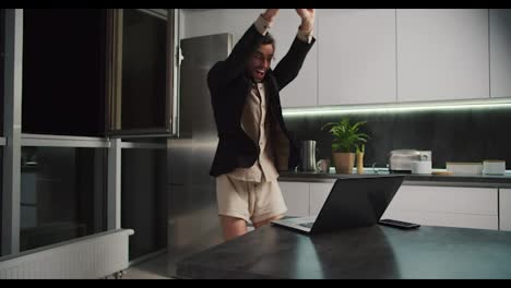 A-brunette-man-with-stubble-in-a-black-jacket-and-beige-shorts-rejoices-at-his-success-and-has-fun-while-working-remotely-in-the-kitchen-in-a-modern-apartment-in-the-evening