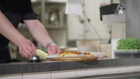 Primer-Plano:-Un-Cocinero-Con-Uniforme-Negro-Coloca-La-Tarta-De-Queso-Terminada-En-Una-Tabla-Sobre-La-Mesa-De-Servicio-Y-Hace-Sonar-La-Campana-Para-Avisar-A-Los-Camareros-Que-El-Plato-Está-Listo.