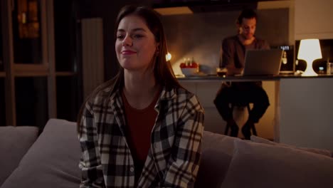 A-calm-and-interested-brunette-girl-in-a-checkered-shirt-sits-on-a-gray-sofa-and-watches-TV-flipping-through-channels-using-the-remote-control-while-her-boyfriend-works-on-a-laptop-in-the-kitchen-in-a-studio-apartment-in-the-evening