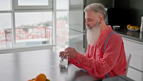 Side-view-of-an-elderly-man-with-gray-hair-and-a-lush-beard-in-a-pink-shirt-with-suspenders-pours-white-medicine-into-a-glass-of-water-and-stirs-it-with-a-spoon-in-a-modern-apartment