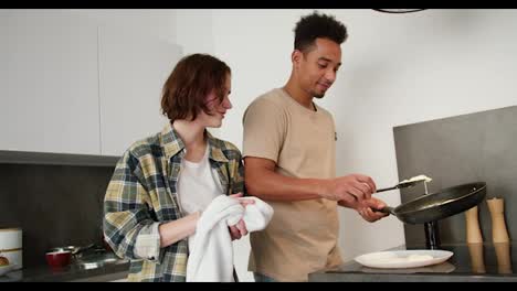 A-happy-young-man-with-Black-skin-in-a-beige-T-shirt-puts-ready-made-scrambled-eggs-on-a-White-plate-he-is-hugged-by-his-young-adult-girl-with-a-bob-hairstyle-in-a-green-shirt-while-preparing-for-breakfast-in-a-modern-kitchen-in-an-apartment