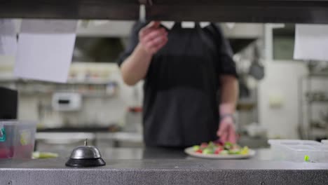 A-professional-male-chef-in-a-black-uniform-places-a-ready-made-salad-order-on-the-serving-table-and-rings-the-bell-to-notify-the-waiters