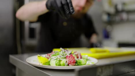 Primer-Plano-De-Un-Cocinero-Confiado-Con-Ropa-Protectora-Negra-Y-Guantes-Protectores-De-Goma-Negros-Rocía-Condimentos-En-Una-Ensalada-Preparada-En-La-Cocina-Antes-De-Servirla-En-Un-Restaurante