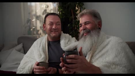 A-happy-elderly-man-with-gray-hair-and-a-lush-gray-beard-is-wrapped-in-a-white-woolen-blanket-and-drinks-hot-tea-with-his-brunette-boyfriend-in-a-gray-gray-T-shirt-on-the-sofa-in-the-evening-in-a-modern-apartment