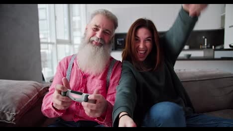 Retrato-De-Un-Anciano-Feliz-Con-Cabello-Gris-Y-Una-Barba-Exuberante-Con-Una-Camisa-Rosa-Que-Juega-Videojuegos-Usando-Joysticks-Con-Su-Hija-Adulta-Morena-Con-Una-Chaqueta-Verde-Oscuro-En-Un-Sofá-Marrón-En-Un-Apartamento-Moderno.