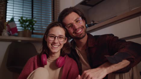 Portrait-of-a-Happy-duo-young-brunette-girl-in-glasses-and-wireless-red-headphones-together-with-her-brunette-boyfriend-with-stubble-sitting-on-the-floor-in-a-modern-apartment-in-the-kitchen-and-posing