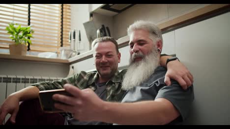 A-happy-elderly-man-with-gray-hair-and-a-lush-gray-beard-in-a-gray-T-shirt-sits-on-the-floor-with-his-brunette-boyfriend-with-stubble-and-a-green-checkered-shirt-and-looks-at-their-photos-on-a-smartphone-in-the-kitchen-during-the-day