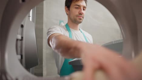 View-from-inside-the-washing-machine-a-confident-brunette-guy-in-a-white-T-shirt-and-a-blue-apron-throws-dirty-things-into-the-washing-machine-and-closes-it