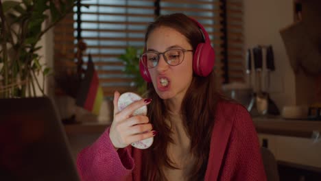 Close-up,-a-young-confident-brunette-girl-with-glasses-in-red-wireless-headphones-and-a-sweater-studies-a-foreign-language-German-and-looks-at-herself-with-the-help-of-a-mirror-while-pronouncing-words-in-a-modern-apartment-in-the-kitchen