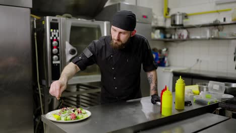 Confident-male-chef-in-a-black-bandana-and-uniform-sprinkles-ready-made-salad-with-sesame-seeds-and-spices-before-serving-in-a-restaurant-kitchen