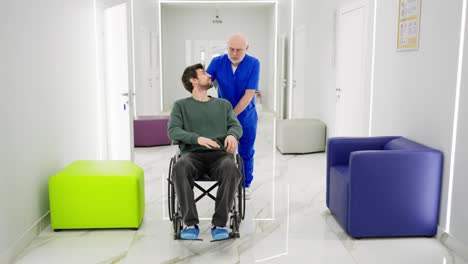 An-experienced-male-doctor-in-glasses-with-a-gray-beard-and-in-a-blue-uniform-communicates-with-a-young-brunette-guy-with-stubble-in-a-green-jacket-who-is-riding-in-a-chair-for-the-disabled-after-his-fracture-during-rehabilitation-in-a-modern-clinic