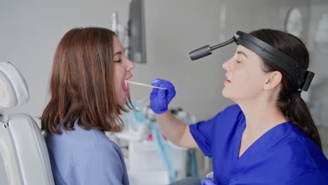 Close-up-of-a-confident-young-brunette-girl-at-an-appointment-with-an-ENT-doctor-in-a-modern-clinic.-A-brunette-doctor-girl-in-a-blue-uniform-puts-a-special-stick-in-the-patients-mouth-to-check-the-throat