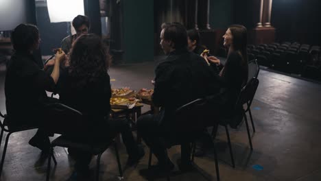 Close-up-over-the-shoulder-of-confident-theater-actors-in-black-suits-sitting-on-chairs-around-and-eating-pizza-during-a-break-and-preparation-for-the-performance-on-stage-in-the-theater