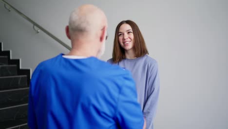 Por-Encima-Del-Hombro,-Una-Chica-Morena-Segura-De-Sí-Misma-Con-Una-Chaqueta-Azul-Se-Comunica-Con-Un-Médico-Experimentado-De-Cabello-Gris-Con-Gafas-Y-Un-Uniforme-Azul-Que-Sostiene-Una-Tableta-Con-Diagnósticos-Y-Consejos-En-Sus-Manos-En-Una-Clínica-Moderna.