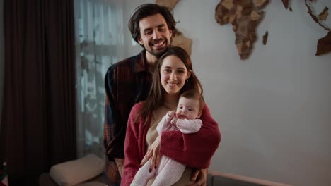 Portrait-of-a-happy-brunette-man-with-stubble-along-with-his-brunette-wife-and-little-baby-daughter-in-a-pink-overalls-posing-in-a-modern-apartment