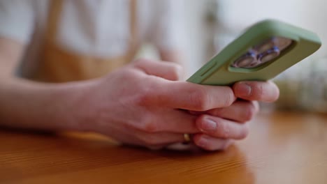 Close-up-a-guy-waiter-in-a-white-T-shirt-and-light-brown-apron-is-scrolling-through-social-networks-on-a-green-smartphone-leaning-on-the-counter-in-a-cafe