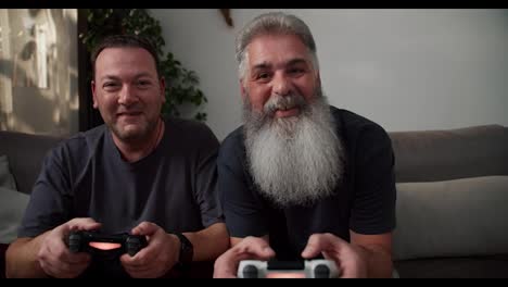 Retrato-De-Un-Hombre-Feliz-Con-Cabello-Gris-Y-Una-Exuberante-Barba-Gris-Jugando-Videojuegos-Usando-Un-Joystick-Junto-Con-Su-Novio-Moreno-Con-Barba-Incipiente-Con-Una-Camiseta-Gris-Sentado-En-El-Sofá-En-Un-Apartamento-Moderno-Por-La-Noche.