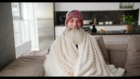 Portrait-of-a-smiling-elderly-man-with-a-lush-gray-beard-who-is-wrapped-in-a-warm-white-blanket-and-sits-in-a-hat-on-a-brown-sofa-in-a-modern-apartment