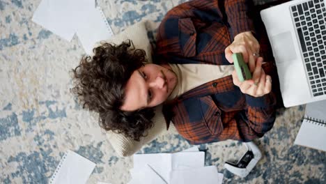 Rotating-camera-happy-brunette-guy-with-curly-hair-lies-on-the-floor-on-a-pillow-and-types-his-ideas-into-a-green-smartphone