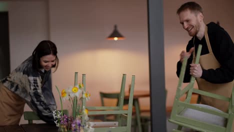 Happy-guy-with-a-beard-in-a-light-apron-lifts-chairs-with-his-colleague-while-cleaning-in-a-cafe.-Teamwork-between-a-guy-and-a-female-waiter-while-cleaning-the-cafe-hall