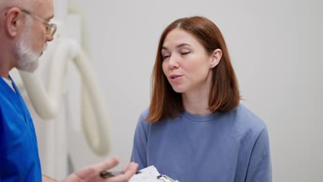 A-confident-brunette-girl-in-a-blue-jacket-communicates-with-an-elderly-doctor-in-a-blue-uniform-during-her-appointment-in-a-modern-clinic