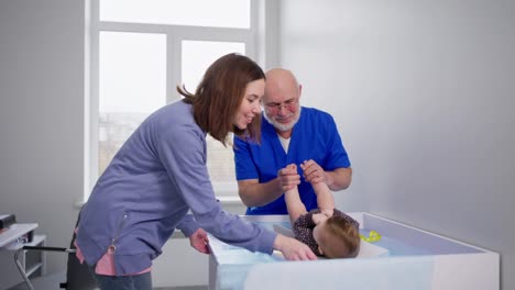 Un-Hombre-Seguro-De-Sí-Mismo,-Con-Barba-Gris-Y-Gafas,-Un-Pediatra-Experimentado-Con-Uniforme-Médico-Azul-Juega-Con-Una-Niña-Bebé-Y-Comprueba-Sus-Normas-De-Desarrollo-A-Su-Edad-En-Una-Clínica-Moderna-Junto-Con-La-Madre-De-La-Niña.