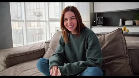 Portrait-of-a-happy-brunette-girl-smiling-widely-in-a-green-sweater-and-jeans-sitting-on-a-modern-brown-sofa-in-the-apartment