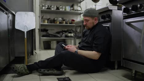 Bored-professional-male-chef-in-a-black-uniform-checks-social-networks-using-his-white-smartphone-while-sitting-on-the-floor-in-the-kitchen-during-a-hard-day-at-work-in-a-restaurant