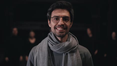 Portrait-of-a-confident-brunette-man-in-round-glasses-stage-director-in-a-gray-T-shirt-and-scarf-whom-actors-in-black-suits-run-up-to-and-hug-from-behind-on-stage-in-the-theater