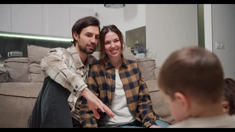 Happy-brunette-man-with-stubble-in-a-checkered-beige-shirt-hugs-his-brunette-wife-in-a-white-t-shirt-and-checkered-shirt-while-they-watch-their-little-children-being-creative-and-drawing-after-moving-to-a-new-apartment-in-a-modern-apartment