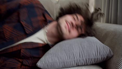 A-happy-brunette-guy-with-curly-hair-in-a-checkered-shirt-lies-down-on-a-soft-pillow-and-with-a-smile-on-his-face-rests-at-home-on-the-sofa-in-a-modern-apartment