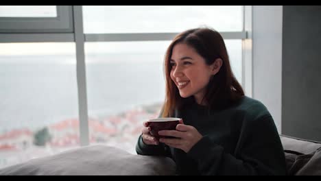 Ein-Glückliches-Brünettes-Mädchen-Freut-Sich,-Während-Es-Auf-Dem-Sofa-Sitzt-Und-Tee-Aus-Einer-Braunen-Tasse-In-Einem-Modernen-Apartment-Mit-Blick-Auf-Das-Meer-Trinkt