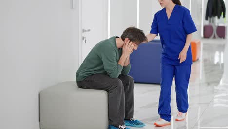 A-sad-and-anxious-brunette-man-in-a-green-jacket-sits-on-a-chair-and-feels-sad-he-needs-to-be-alone-during-a-serious-diagnosis-in-a-modern-clinic.-A-brunette-doctor-girl-in-a-blue-uniform-tries-to-support-the-guy-but-she-hardly-succeeds