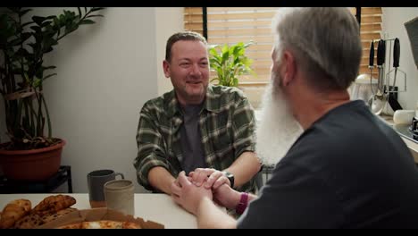 Over-the-shoulder-a-happy-brunette-man-with-stubble-in-a-green-plaid-shirt-holds-hands-with-his-older-boyfriend-with-gray-hair-and-a-lush-gray-beard-in-a-gray-T-shirt-during-his-conversation-in-the-kitchen-at-the-table