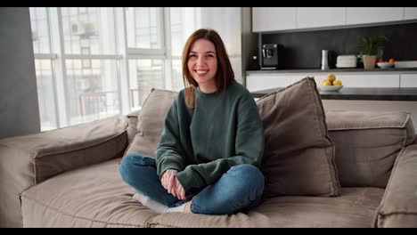 Portrait-of-a-happy-brunette-girl-in-a-dark-green-sweater-and-jeans-who-is-sitting-on-a-modern-brown-sofa-in-a-studio-apartment