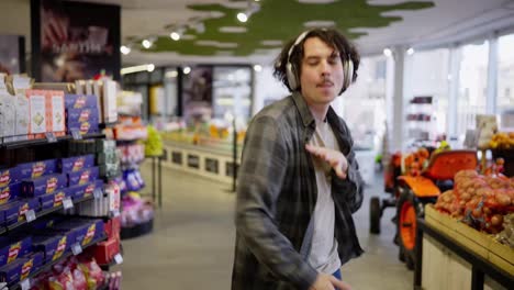 Joyful-brunette-guy-in-a-checkered-shirt-with-white-headphones-listens-to-music-walks-and-dances-intensely-in-a-grocery-store-while-shopping