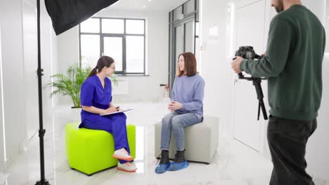 Ein-Selbstbewusstes-Brünettes-Mädchen-In-Einem-Blauen-Pullover-Interviewt-Eine-Brünette-Ärztin-Mit-Brille-In-Einer-Herbstuniform-In-Einer-Modernen,-Hellen-Klinik.-Dies-Wird-Von-Einem-Pressevertreter-Gefilmt.