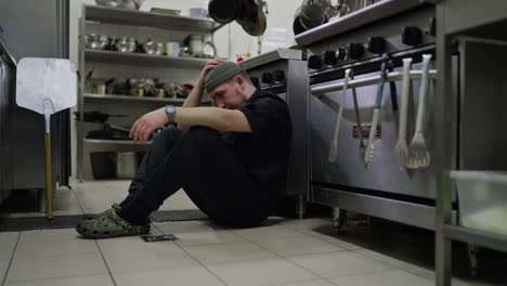 A-tired-male-professional-chef-in-a-black-uniform-sits-on-the-floor-in-the-kitchen-leaning-on-the-table-and-tries-to-relax-and-collect-his-thoughts-during-a-hard-busy-day-in-the-kitchen-of-a-restaurant