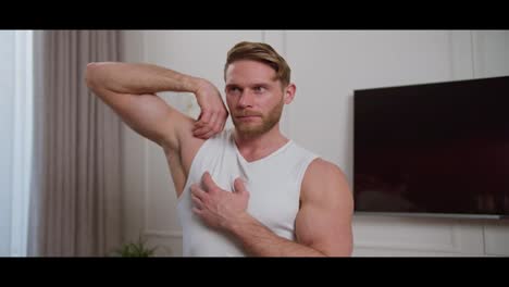 Confident-blond-athletic-man-with-stubble-in-a-white-T-shirt-doing-arm-swings-and-stretching-his-shoulders-before-starting-a-workout-at-home-in-a-modern-apartment-with-a-white-wall