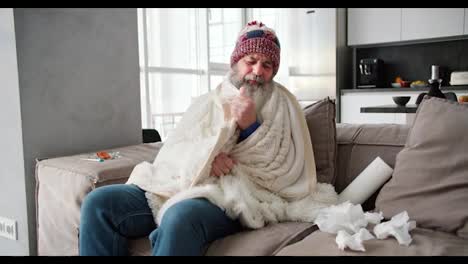 Portrait-of-a-sick-elderly-man-with-a-lush-beard-and-a-red-hat-wrapped-in-a-white-blanket-and-folds-used-napkins-near-him-while-sitting-on-the-sofa-near-a-thermometer-and-a-set-of-medications-in-a-modern-apartment-on-the-sofa