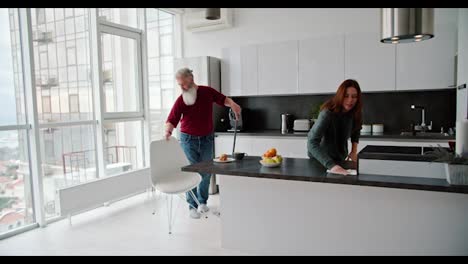 An-elderly-man-with-gray-hair-and-a-lush-beard-in-a-red-T-shirt-is-cleaning-his-home-with-his-adult-brunette-daughter-in-a-green-sweater-in-a-modern-kitchen