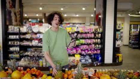 Happy-brunette-guy-with-curly-hair-shows-various-fruits-while-blogging-using-a-smartphone-in-a-supermarket