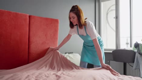 A-brunette-cleaning-lady-girl-in-a-white-T-shirt-and-blue-apron-makes-the-bed-with-a-pink-blanket-and-gray-pillows-while-cleaning-the-bedroom-when-the-cleaning-company-was-called