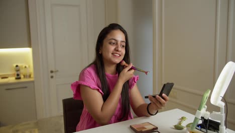 A-happy-Indian-brunette-girl-in-a-pink-dress-sits-in-front-of-the-lighting-and-her-green-smartphone-while-blogging-online-about-how-to-properly-apply-cosmetics-and-do-Makeup-in-a-modern-apartment-during-the-day