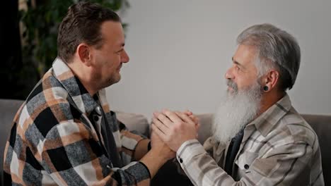 From-the-side,-a-happy-brunette-with-stubble-in-a-plaid-shirt-holds-the-hands-of-his-elderly-boyfriend-with-gray-hair-and-a-full-beard,-and-they-press-their-foreheads-to-each-other,-looking-into-the-eyes-while-sitting-on-the-sofa-in-a-modern-apartment