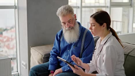 Eine-Brünette-Frau-In-Einem-Weißen-Arztkittel-Zeigt-Einem-Mann-Mit-Grauem-Haar,-Brille-Und-üppigem-Bart-Auf-Einem-Tablet-Ihre-Namen-Und-Rezepte-Für-Medikamente-Zur-Verbesserung-Der-Gesundheit-Nach-Einer-Hausuntersuchung-Bei-Einem-Arzt-In-Einer-Modernen-Wohnung