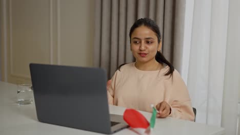 Una-Feliz-Chica-India-Morena-Con-Un-Suéter-Beige-Estudia-Idiomas-Extranjeros,-Italiano-En-Línea-Y-Se-Comunica-Con-El-Profesor-A-Través-De-Una-Videoconferencia-En-Computadoras-Portátiles-Grises-En-Un-Apartamento-Moderno-Durante-El-Día