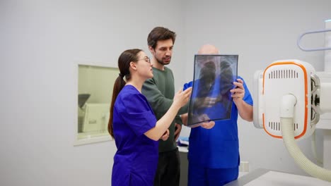 Un-Hombre-Moreno-Con-Barba-Incipiente-Y-Chaqueta-Verde-Se-Comunica-Con-Una-Niña-Y-Un-Médico-Con-Uniforme-Azul-Después-De-Realizar-Una-Fluorografía-Frente-A-Una-Imagen-De-Su-Propio-Pecho-En-Una-Hoja-Transparente-Especial-En-Una-Oficina-De-Una-Clínica-Moderna.