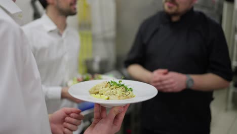 Primer-Plano:-Un-Camarero-Con-Una-Camisa-Blanca-Sostiene-Un-Plato-Preparado-En-Sus-Manos-Y-Se-Comunica-Con-Un-Chef-Profesional-Con-Un-Uniforme-Negro-Sobre-Servirlo-Junto-Con-Su-Colega-Camarero-En-La-Cocina-De-Un-Restaurante.