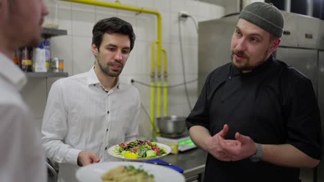 Close-up-over-the-shoulder-of-a-confident-male-chef-in-a-black-uniform-talking-with-waiters-about-teamwork-in-the-kitchen-in-a-restaurant.-A-professional-chef-is-dissatisfied-with-the-work-of-waiters-and-communicates-with-them-to-solve-professional-problems